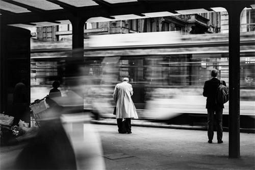 alt="Blurred tram passing by a station with waiting passengers"